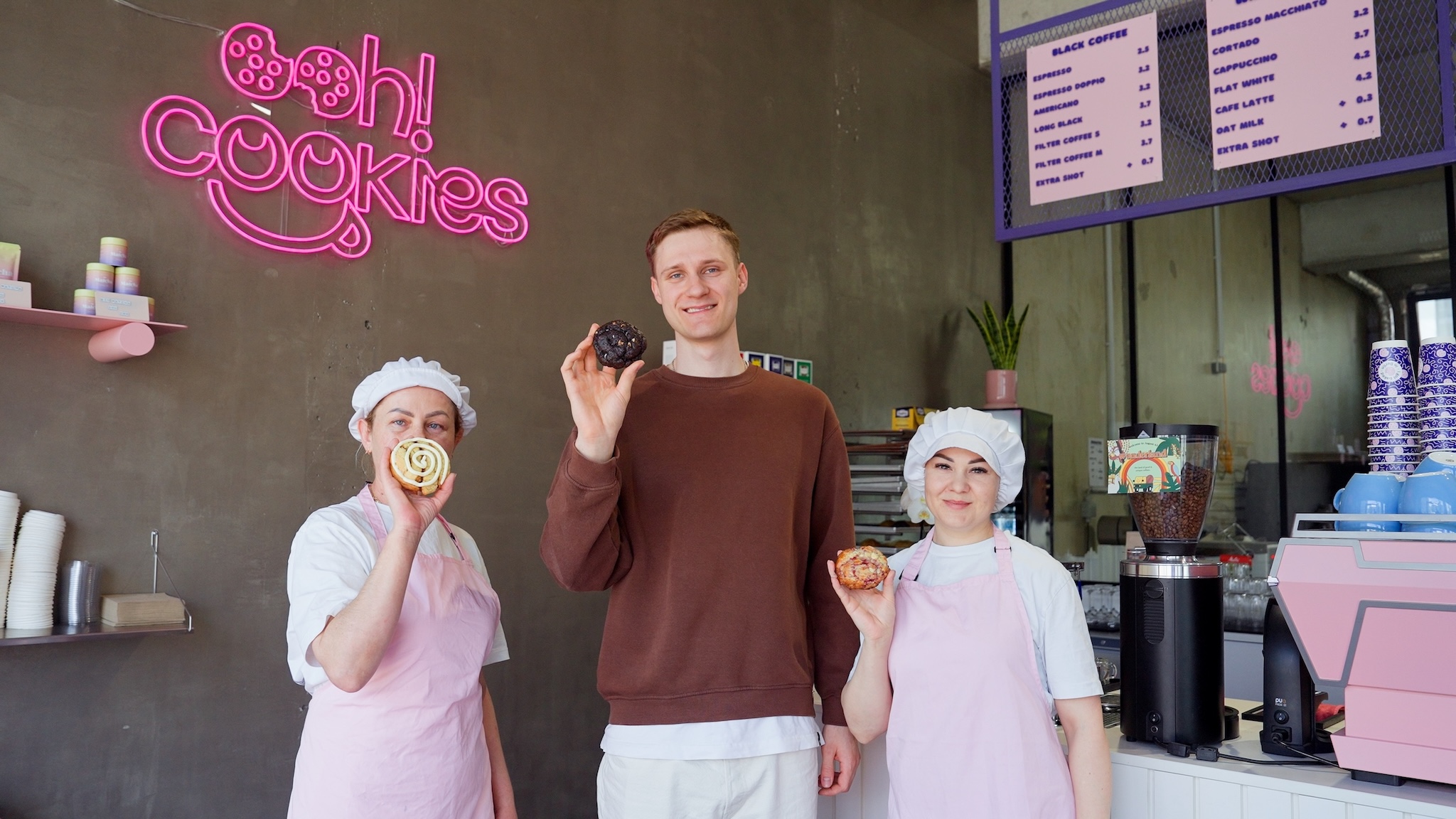 Inhaber Max Hartfiel und zwei Mitarbeiterinnen von OOH! Cookies vor dem Neon-Logo des Unternehmens.